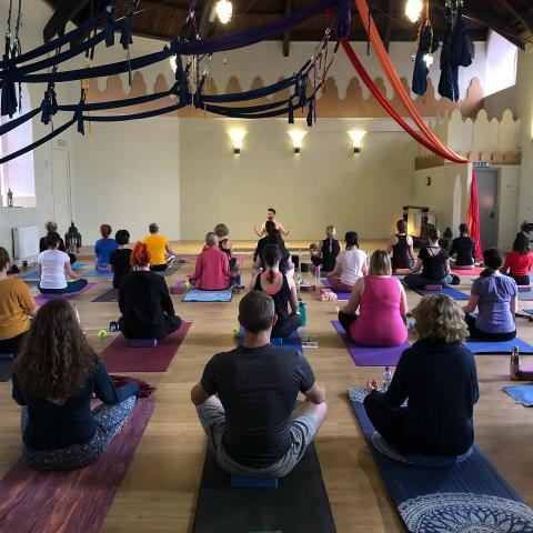 People gathered in a Yoga studio, sitting crossed-legged, looking towards the instructor