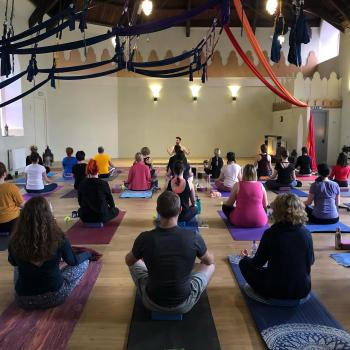 People gathered in a Yoga studio, sitting crossed-legged, looking towards the instructor